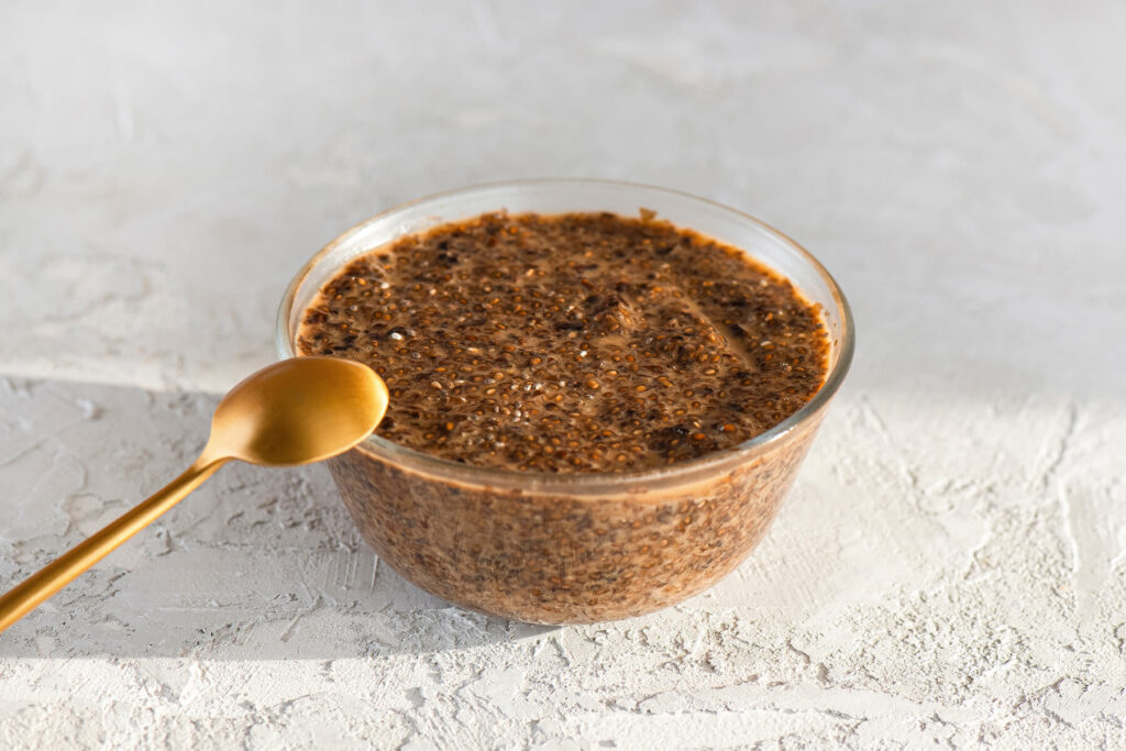 Glasschale mit veganem Chia-Kaviar aus Chiasamen und Sojasauce, dunkelbraune, glänzende Chia-Perlen, ein goldener Löffel lehnt links an der Schale auf hellem Untergrund