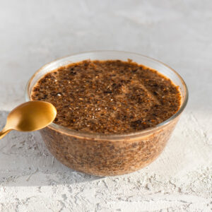 Glasschale mit veganem Chia-Kaviar aus Chiasamen und Sojasauce, dunkelbraune, glänzende Chia-Perlen, ein goldener Löffel lehnt links an der Schale auf hellem Untergrund