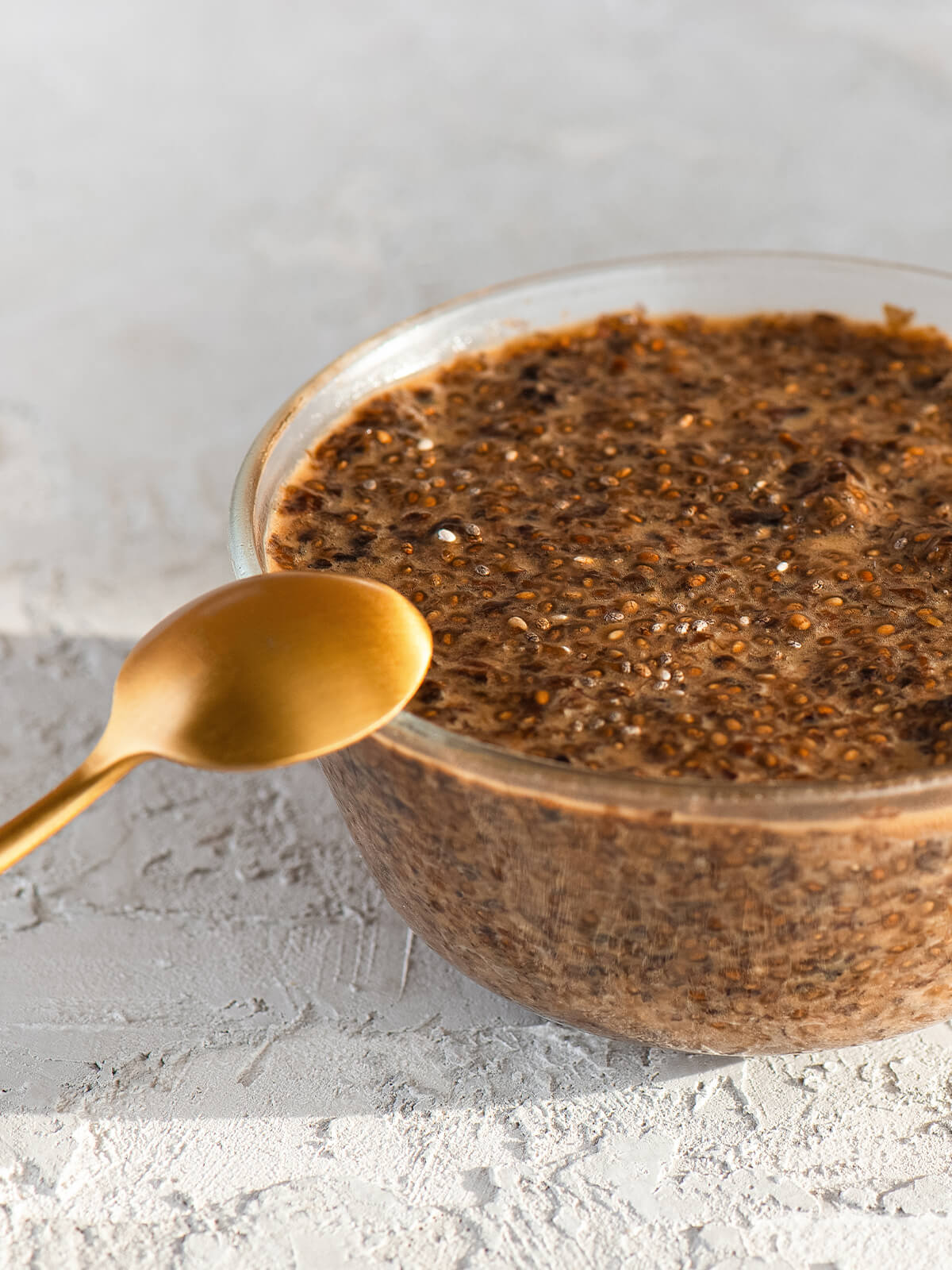 Runde Glasschale mit veganem Chia-Kaviar aus Chiasamen und Sojasauce, dunkelbraune glänzende Perlen, ein goldener Löffel lehnt an der Schale auf hellem, strukturiertem Untergrund