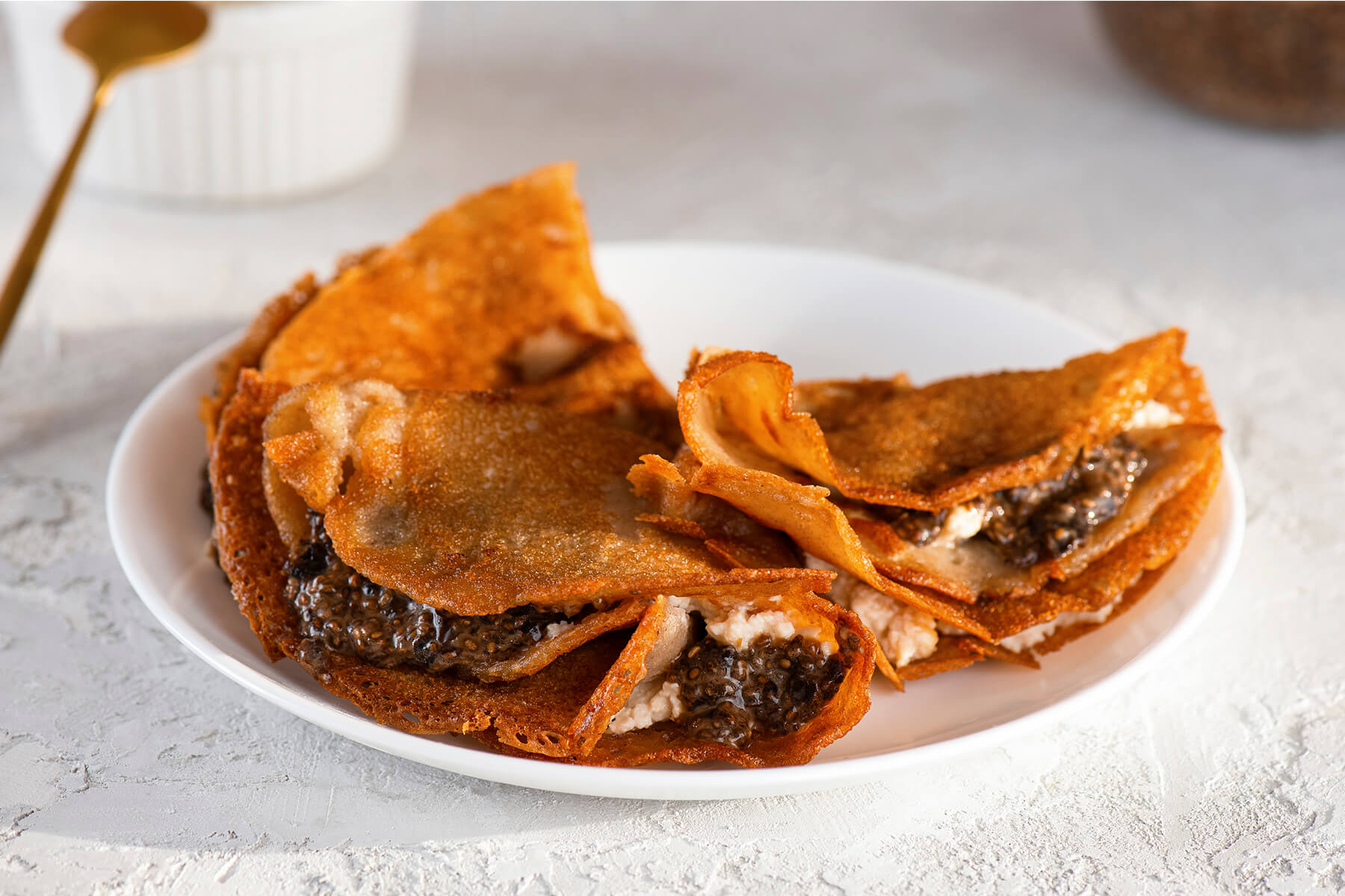 Goldbraune, dünne Crêpes auf einem weißen Teller, gefaltet und gefüllt mit dunklem veganem Chia-Kaviar und weißer veganer Frischkäse-Creme, als Serviervorschlag angerichtet