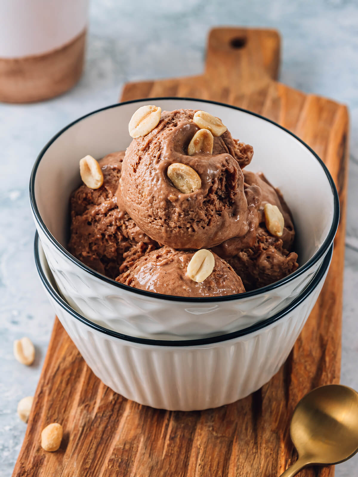 Schoko-Erdnuss-Bananeneis als hohe, milchschokoladenbraune Kugeln in einer weißen Schüssel auf einem Holzbrett, mit hellen blanchierten Erdnusskernen bestreut, daneben ein goldener Löffel und einzelne Kerne
