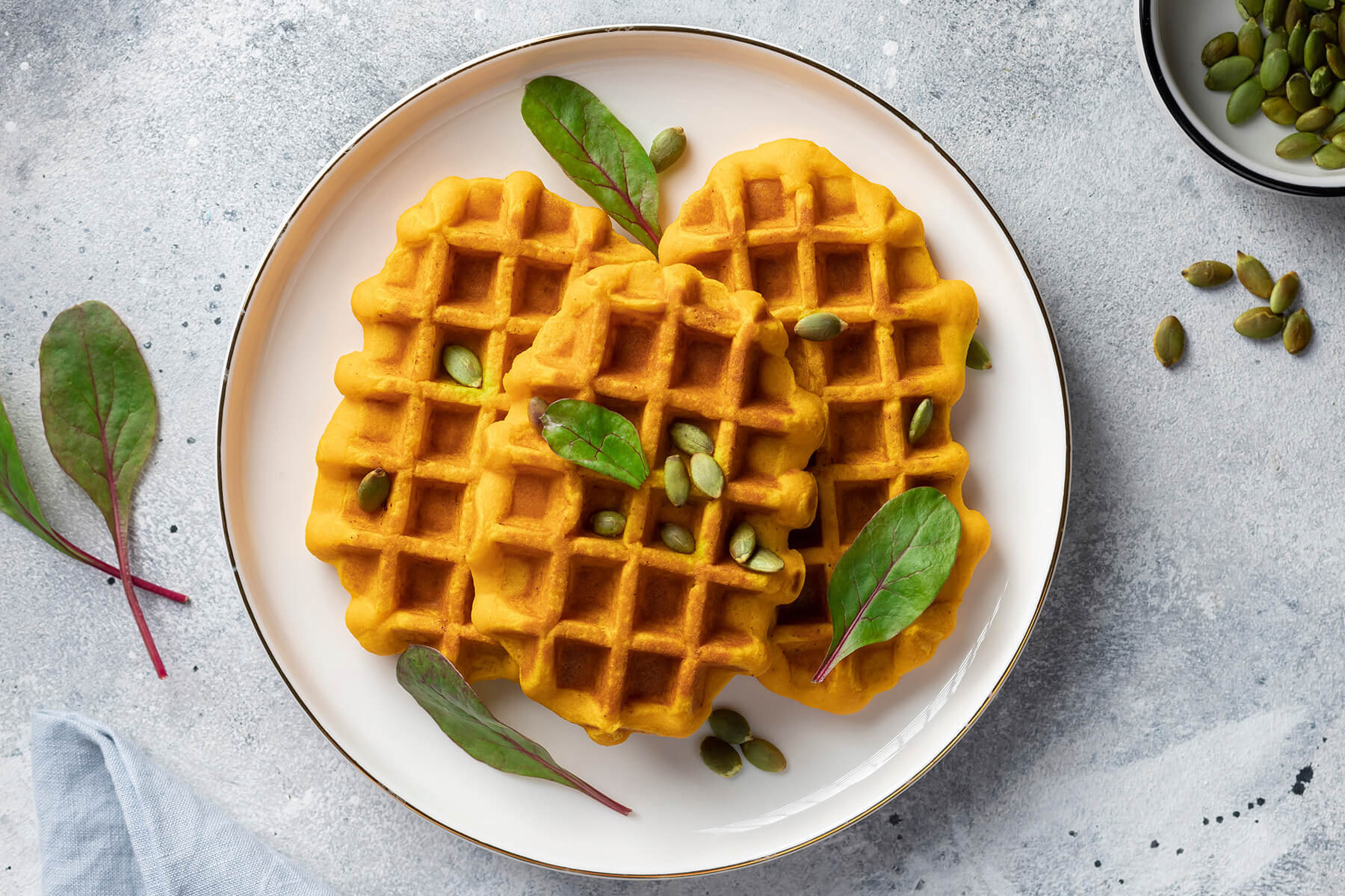 Vegane Kürbiswaffeln auf einem Teller, garniert mit Kürbiskernen und frischen Blättern, daneben eine Schale Kürbiskerne