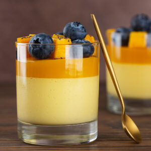 Zwei Gläser vegane Mango-Kokos-Panna-Cotta mit cremig-hellgelber Mango-Kokos-Schicht unten und leuchtend oranger Mangoschicht oben, garniert mit frischen Mangowürfeln, Heidelbeeren und Chiasamen; ein goldener Löffel lehnt am vorderen Glas, dahinter ein dunkler Holztisch und warmer brauner Hintergrund