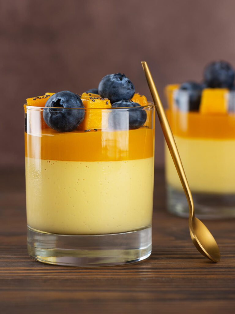 Zwei Gläser vegane Mango-Kokos-Panna-Cotta mit cremig-hellgelber Mango-Kokos-Schicht unten und leuchtend oranger Mangoschicht oben, garniert mit frischen Mangowürfeln, Heidelbeeren und Chiasamen; ein goldener Löffel lehnt am vorderen Glas, dahinter ein dunkler Holztisch und warmer brauner Hintergrund