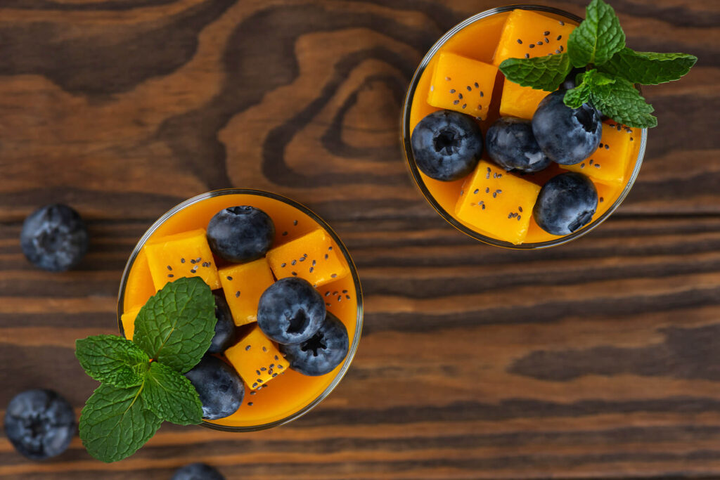 Vegane Mango-Kokos-Panna-Cotta von oben in zwei Gläsern, die orange Mangoschicht dicht belegt mit Mangowürfeln, frischen Heidelbeeren, Chiasamen und einem Minzzweig, auf einem dunklen Holztisch mit einzelnen Heidelbeeren daneben