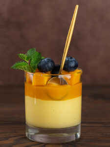 Ein Glas vegane Mango-Kokos-Panna-Cotta mit klar getrennten Schichten, cremig-hellgelber Mango-Kokos-Schicht unten und oranger Mangoschicht oben, garniert mit Mangowürfeln, Heidelbeeren, Chiasamen und einem Minzzweig, ein goldener Löffel steckt im Glas, warmer brauner Hintergrund