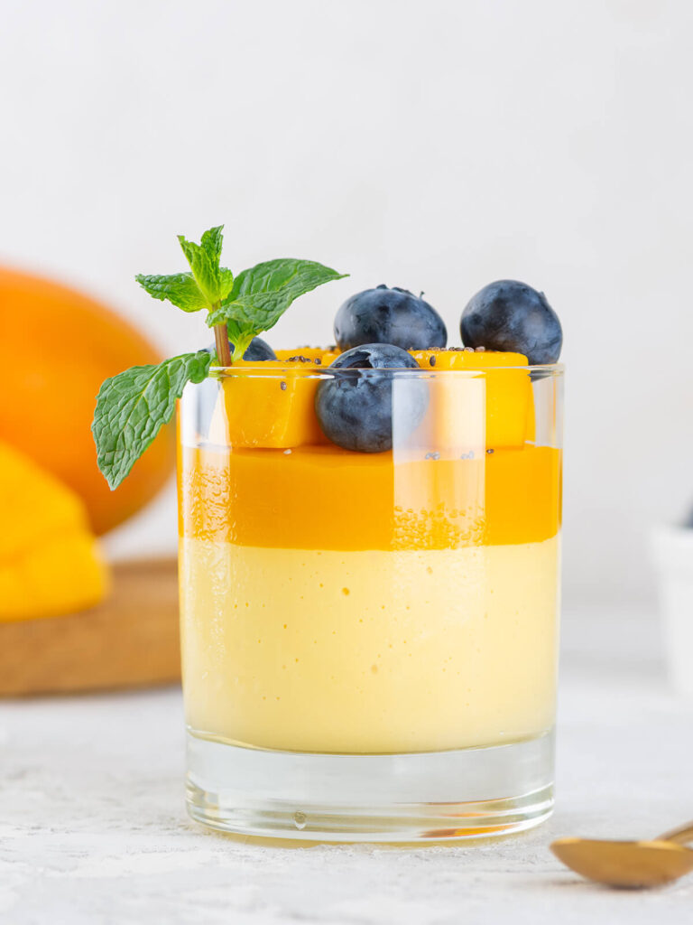 Ein Glas vegane Mango-Kokos-Panna-Cotta vor hellem Hintergrund, unten die cremige hellgelbe Mango-Kokos-Schicht, darüber die orange Mangoschicht, garniert mit Mangowürfeln, Heidelbeeren, Chiasamen und einem Minzzweig, im Hintergrund eine ganze Mango auf einem Holzbrett