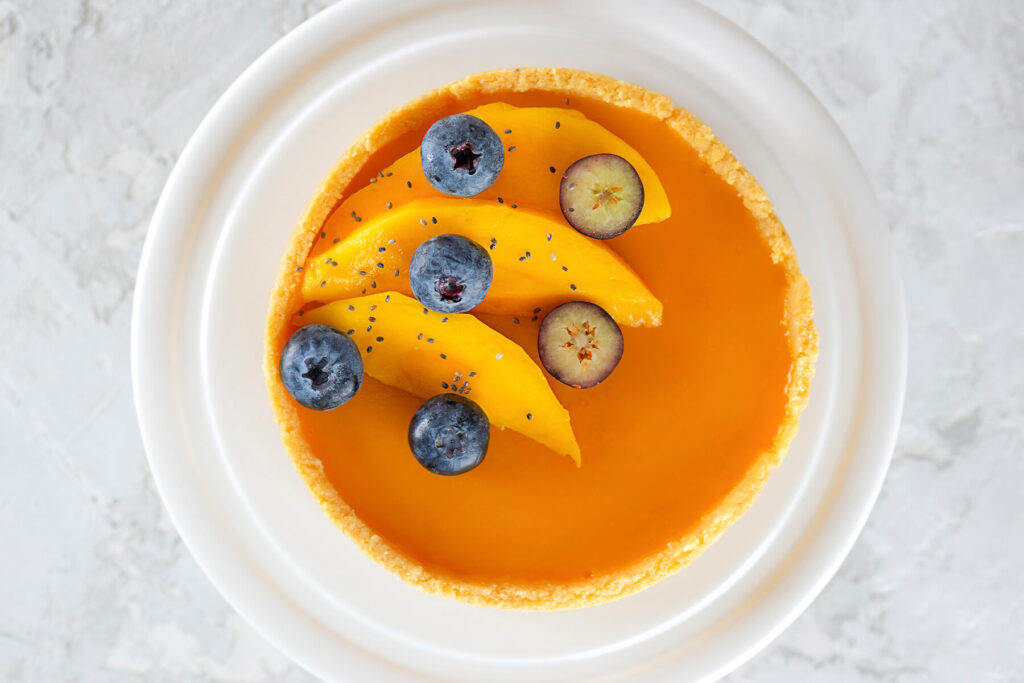 Aufsicht auf eine runde vegane Mango-Tarte auf weißem Teller mit glänzendem orangem Mango-Spiegel und goldenem Mürbeteig-Rand, garniert mit drei Mangospalten, ganzen und quer halbierten Heidelbeeren und wenigen Chiasamen