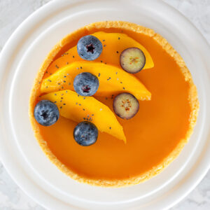 Aufsicht auf eine runde vegane Mango-Tarte auf weißem Teller mit glänzendem orangem Mango-Spiegel und goldenem Mürbeteig-Rand, garniert mit drei Mangospalten, ganzen und quer halbierten Heidelbeeren und wenigen Chiasamen