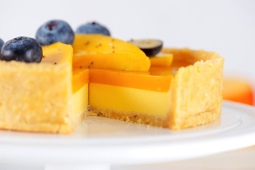 Nahaufnahme einer angeschnittenen veganen Mango-Tarte im Anschnitt auf weißem Teller, deutlich sichtbar der goldene Mürbeteig-Boden, die cremige gelbe Mango-Kokos-Schicht und der orange Mango-Spiegel, garniert mit Mangoscheiben, Heidelbeeren, Chiasamen und einer halbierten Weintraube