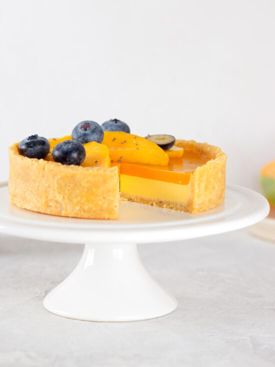 Ganze vegane Mango-Tarte auf einem weißen Tortenständer, ein Stück ist herausgeschnitten und gibt den Blick auf die Schichten aus goldenem Mürbeteig-Boden, gelber Mango-Kokos-Creme und orangem Mango-Spiegel frei, oben mit Mangoscheiben, Heidelbeeren, Chiasamen und einer halbierten Weintraube garniert