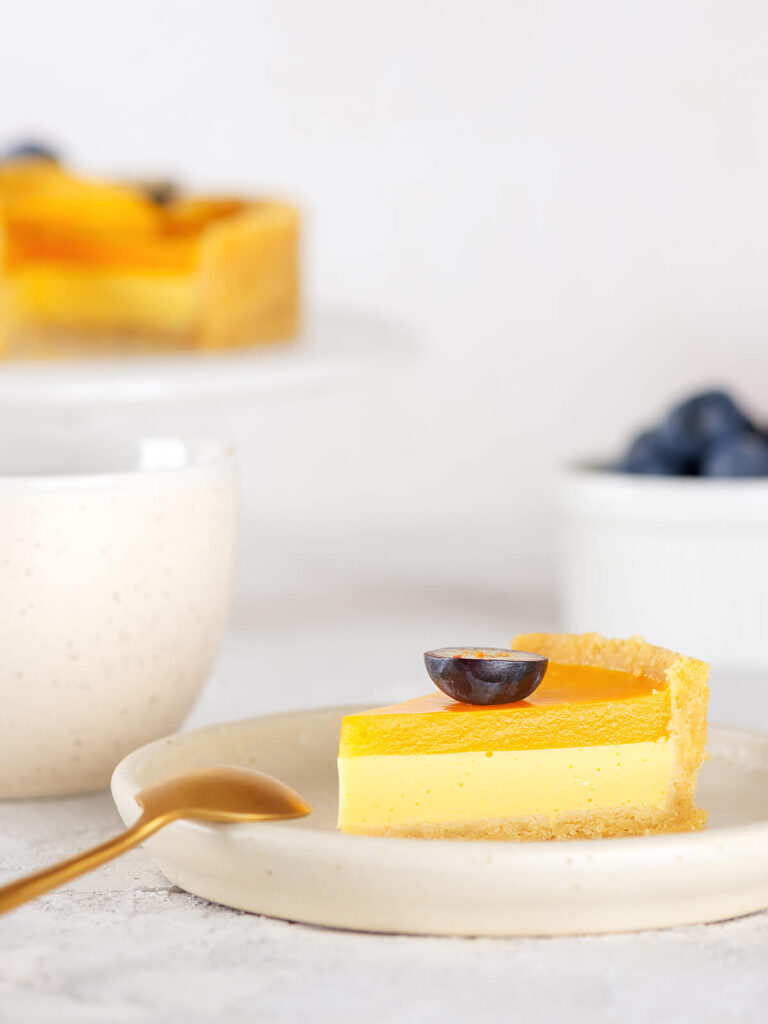 Ein Stück vegane Mango-Tarte auf beigem Teller mit goldenem Löffel und heller Teetasse, im Anschnitt der goldene Mürbeteig-Boden, die gelbe Mango-Kokos-Creme und der orange Mango-Spiegel, oben mit einer quer halbierten Heidelbeere garniert