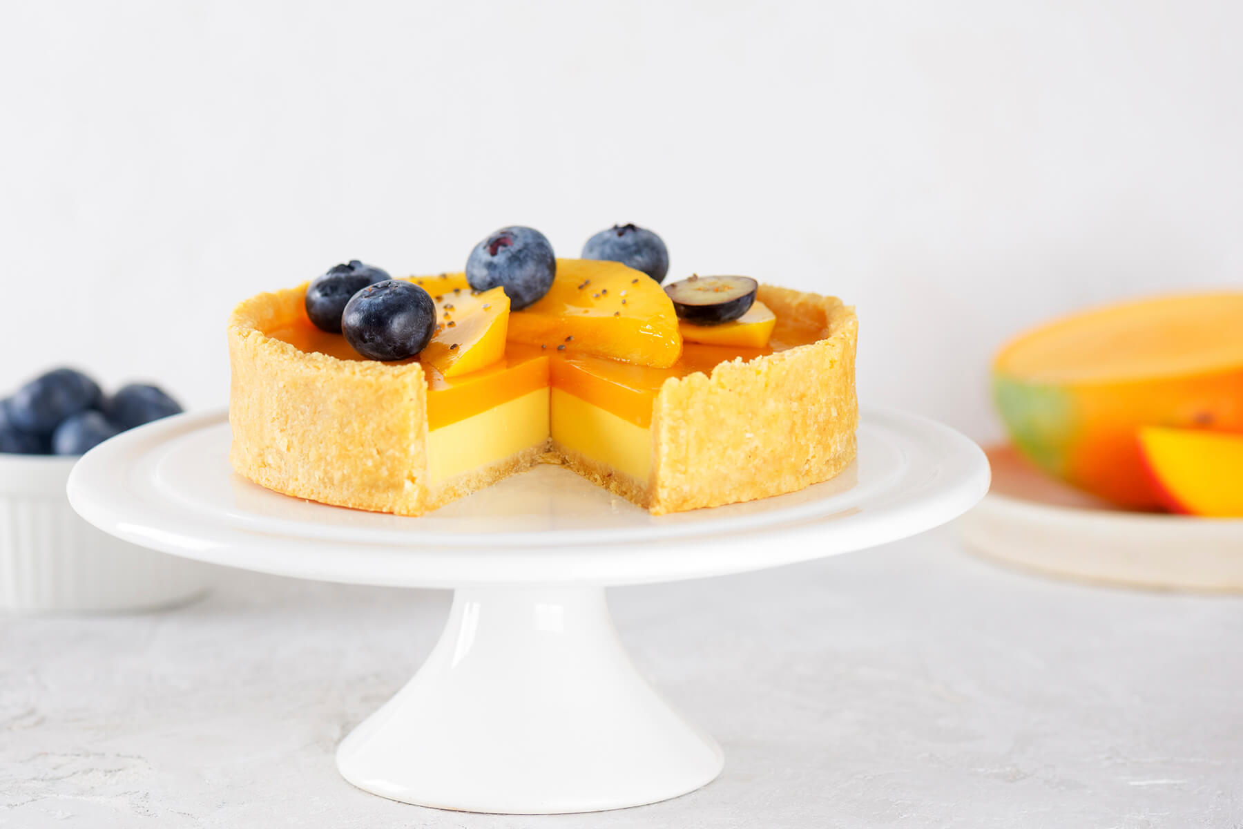 Vegane Mango-Tarte auf weißem Tortenständer mit herausgeschnittenem Stück, im Hintergrund eine aufgeschnittene frische Mango, die Tarte dekoriert mit Heidelbeeren, Mangospalten, Chiasamen und einer quer halbierten Heidelbeere