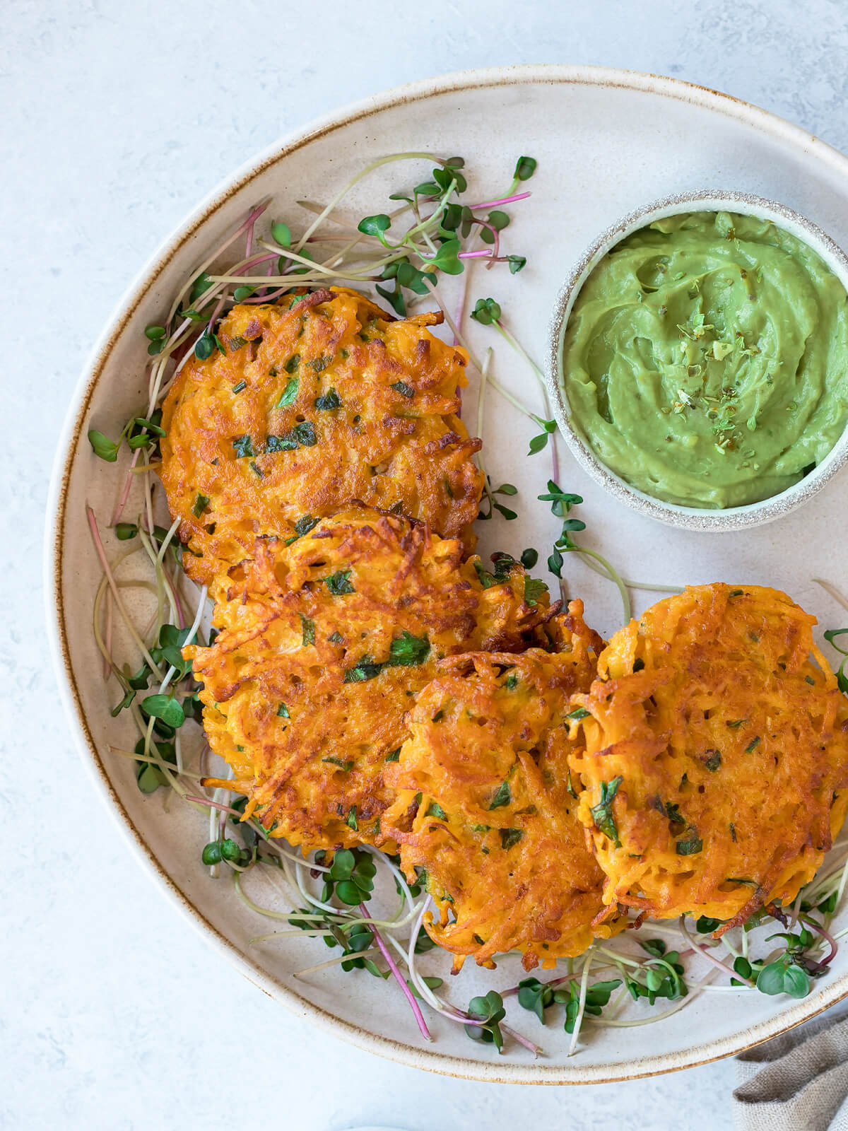 Vegane Süßkartoffel-Puffer mit cremigem Avocado-Dip in einer Schale und frischer Kresse auf einem Teller