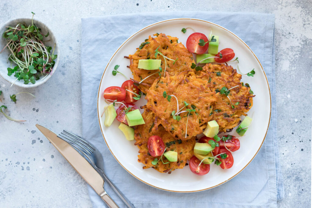 Vegane Süßkartoffel-Puffer mit gewürfelter Avocado, halbierten Kirschtomaten und Kresse auf einem Teller
