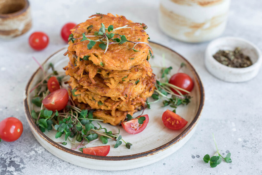 Gestapelte vegane Süßkartoffel-Puffer mit halbierten Kirschtomaten und frischer Kresse auf einem Teller