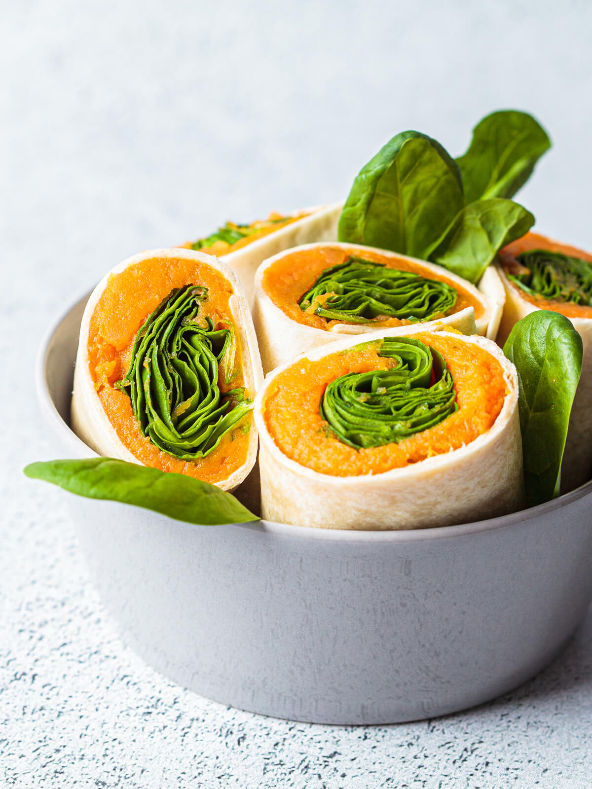 Vegane Wrap-Röllchen mit orangefarbenem Süßkartoffelmus und grünem Spinat in einer grauen Schale, garniert mit frischen Spinatblättern