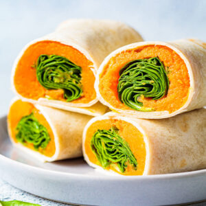 Aufgeschnittene vegane Wrap-Röllchen mit orangefarbenem Süßkartoffelmus und grünem Spinat, gestapelt auf einem hellen Teller