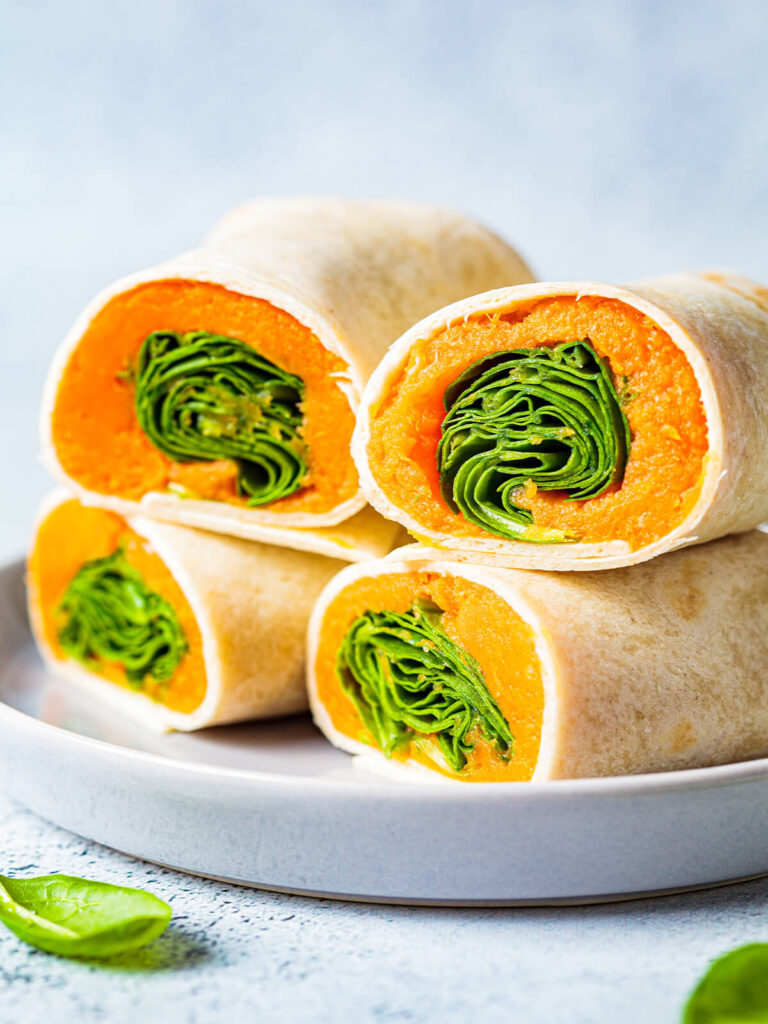 Aufgeschnittene vegane Wrap-Röllchen mit orangefarbenem Süßkartoffelmus und grünem Spinat, gestapelt auf einem hellen Teller