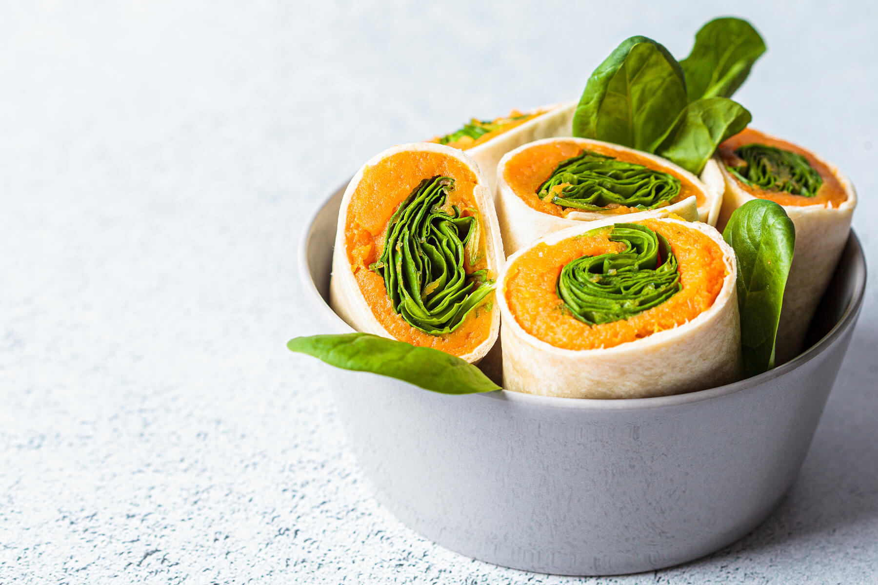 Vegane Süßkartoffel-Spinat-Wraps in Scheiben in einer grauen Schale, daneben frische Spinatblätter auf hellem Untergrund
