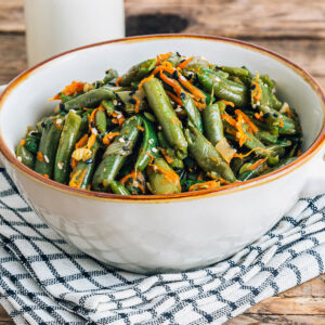 Warmer Bohnen-Karotten-Salat mit Sesam in einer weißen Schüssel mit braunem Rand auf einem karierten Tuch, mit glänzenden grünen Bohnenstücken, geraspelten Möhren sowie schwarzem und weißem Sesam bestreut