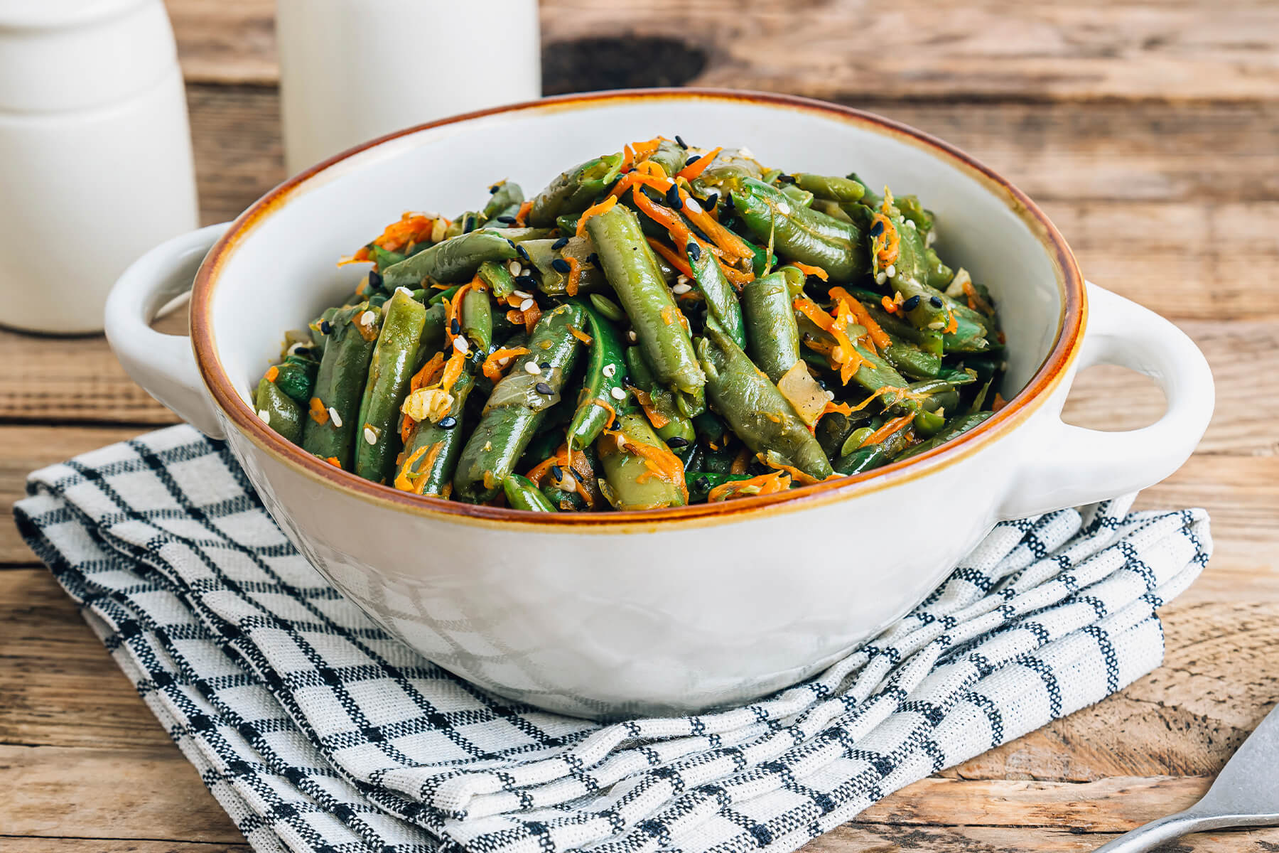 Warmer Bohnen-Karotten-Salat mit Sesam in einer weißen Schüssel mit braunem Rand auf einem karierten Tuch, mit glänzenden grünen Bohnenstücken, geraspelten Möhren sowie schwarzem und weißem Sesam