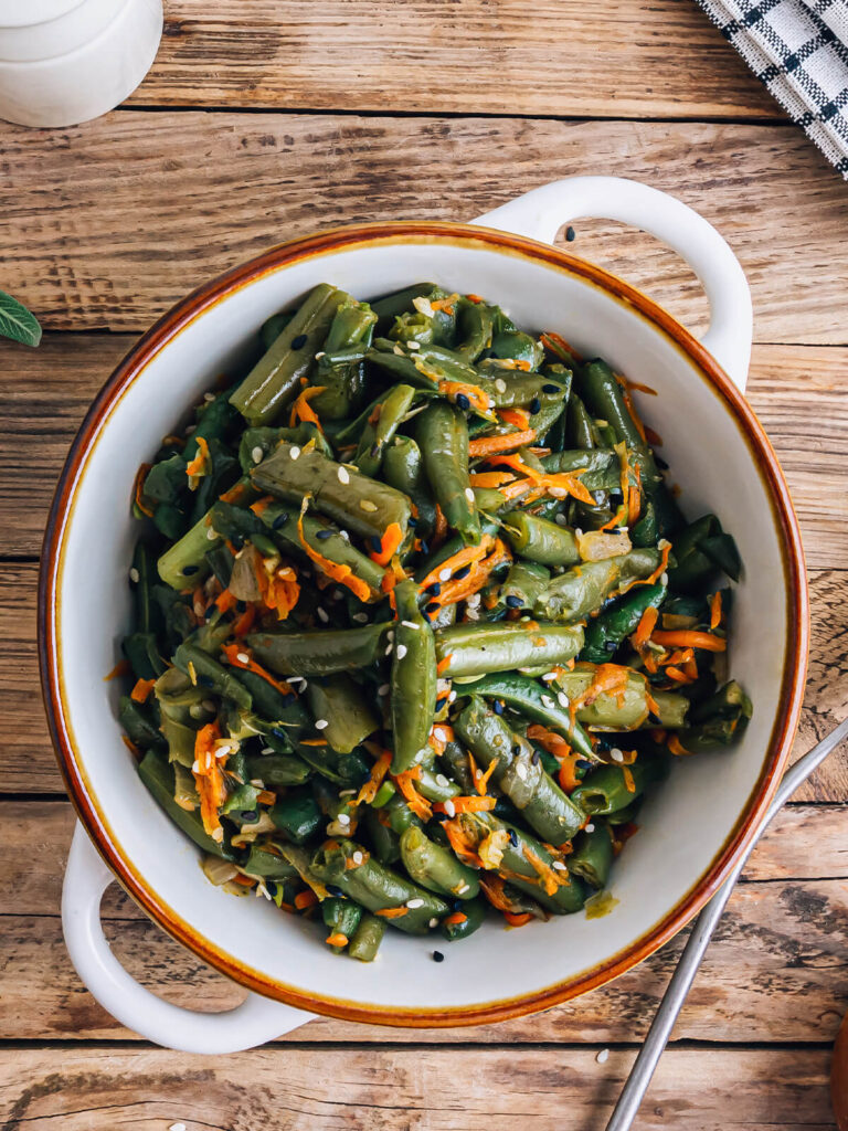 Fertiger warmer Bohnen-Karotten-Salat mit Sesam von oben in einer ovalen weißen Form, mit glänzenden grünen Bohnenstücken, geraspelten Möhren sowie schwarzem und weißem Sesam bestreut