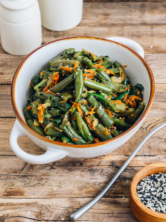 Warmer Bohnen-Karotten-Salat mit Sesam im Hochformat, schräg von der Seite in einer weißen Schüssel mit braunem Rand, mit glänzenden grünen Bohnenstücken, geraspelten Möhren sowie schwarzem und weißem Sesam bestreut, daneben ein Löffel und ein Schälchen mit Sesam