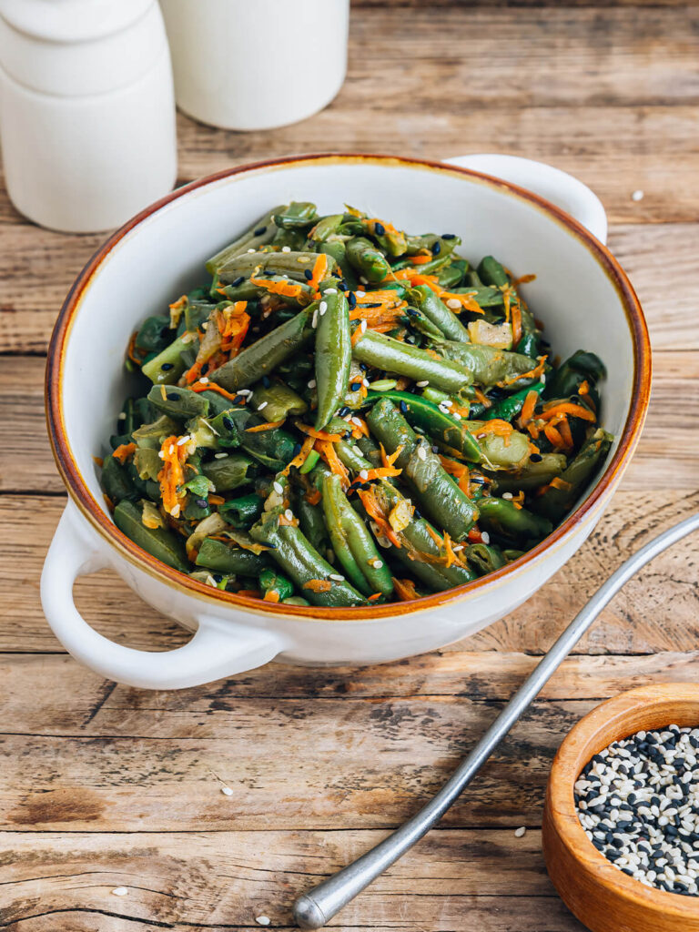 Warmer Bohnen-Karotten-Salat mit Sesam im Hochformat, schräg von der Seite in einer weißen Schüssel mit braunem Rand, mit glänzenden grünen Bohnenstücken, geraspelten Möhren sowie schwarzem und weißem Sesam bestreut, daneben ein Löffel und ein Schälchen mit Sesam