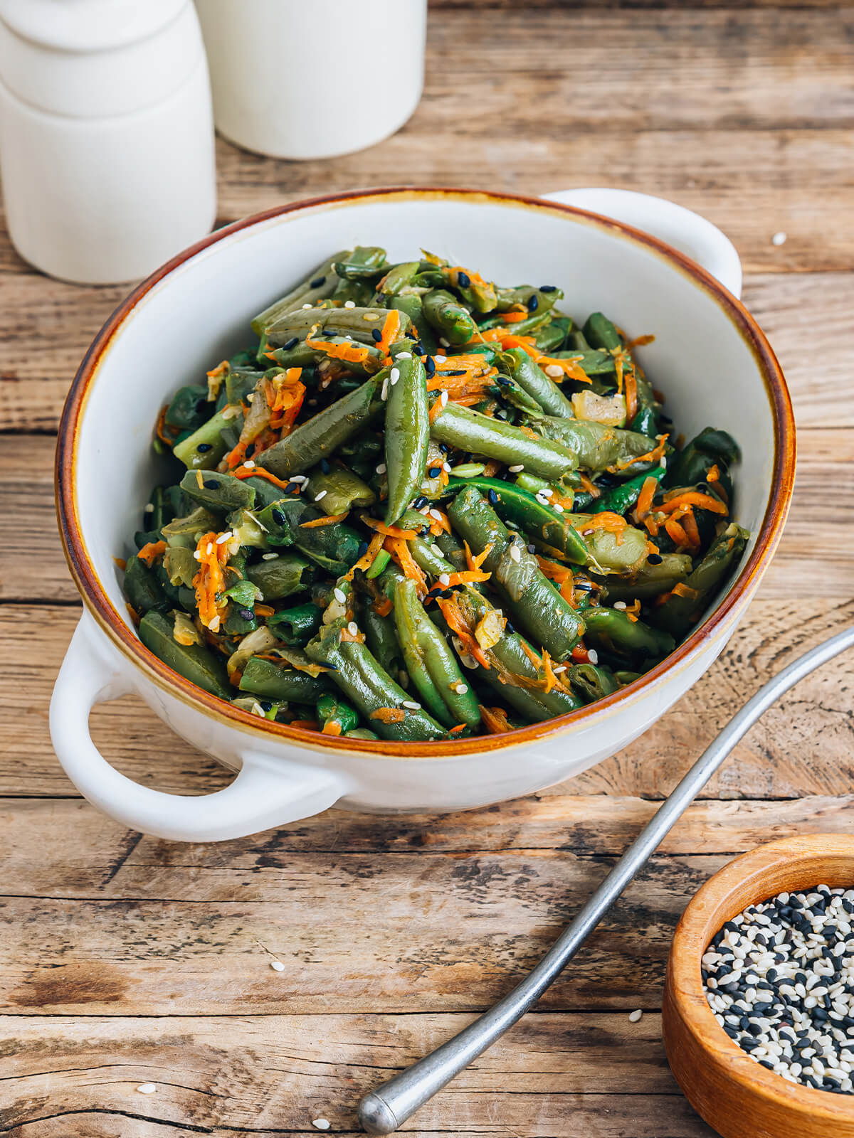 Warmer Bohnen-Karotten-Salat mit Sesam im Hochformat, schräg von der Seite in einer weißen Schüssel mit braunem Rand, mit glänzenden grünen Bohnenstücken, geraspelten Möhren sowie schwarzem und weißem Sesam bestreut, daneben ein Löffel und ein Schälchen mit Sesam