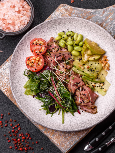 Warmer Roastbeef-Salat mit rosa gegartem Rindfleisch, geschwenkten Blattsalaten, warmen Gurkenbändern, Zucchini, Edamame und Tomatenhälften auf hellem Teller, daneben Salz und rosa Pfeffer