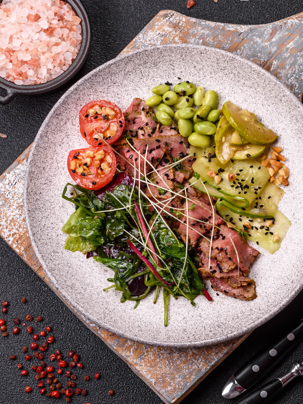 Warmer Roastbeef-Salat mit rosa gegartem Rindfleisch, geschwenkten Blattsalaten, warmen Gurkenbändern, Zucchini, Edamame und Tomatenhälften auf hellem Teller, daneben Salz und rosa Pfeffer