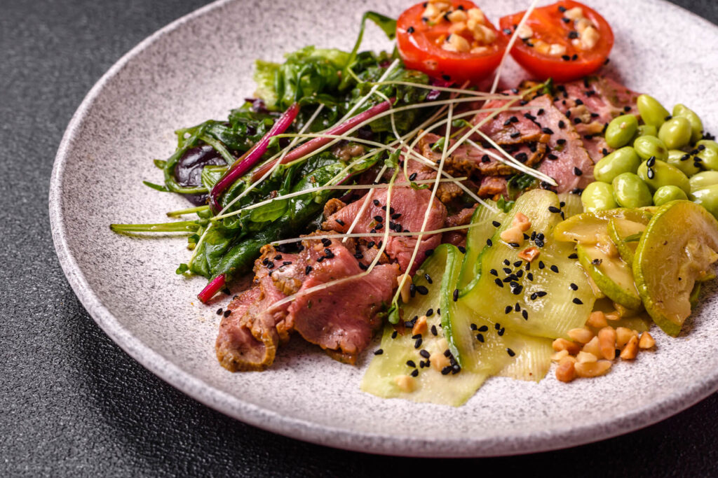 Warmer Roastbeef-Salat mit rosa gegartem Rindfleisch, geschwenkten Blattsalaten, warmen Gurkenbändern, Zucchini, Edamame, Tomatenhälften und schwarzem Sesam auf hellem Teller