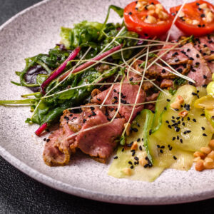 Warmer Roastbeef-Salat mit rosa gegartem Rindfleisch, geschwenkten Blattsalaten, warmen Gurkenbändern, Zucchini, Edamame, Tomatenhälften und schwarzem Sesam auf hellem Teller
