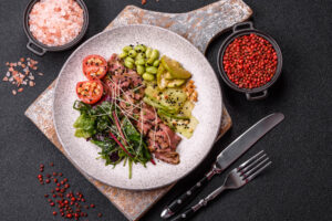 Warmer Roastbeef-Salat von oben auf einem Holzbrett, mit geschwenkten Blattsalaten, Edamame, warmen Gurkenbändern und Tomatenhälften, daneben Salz und rosa Pfeffer