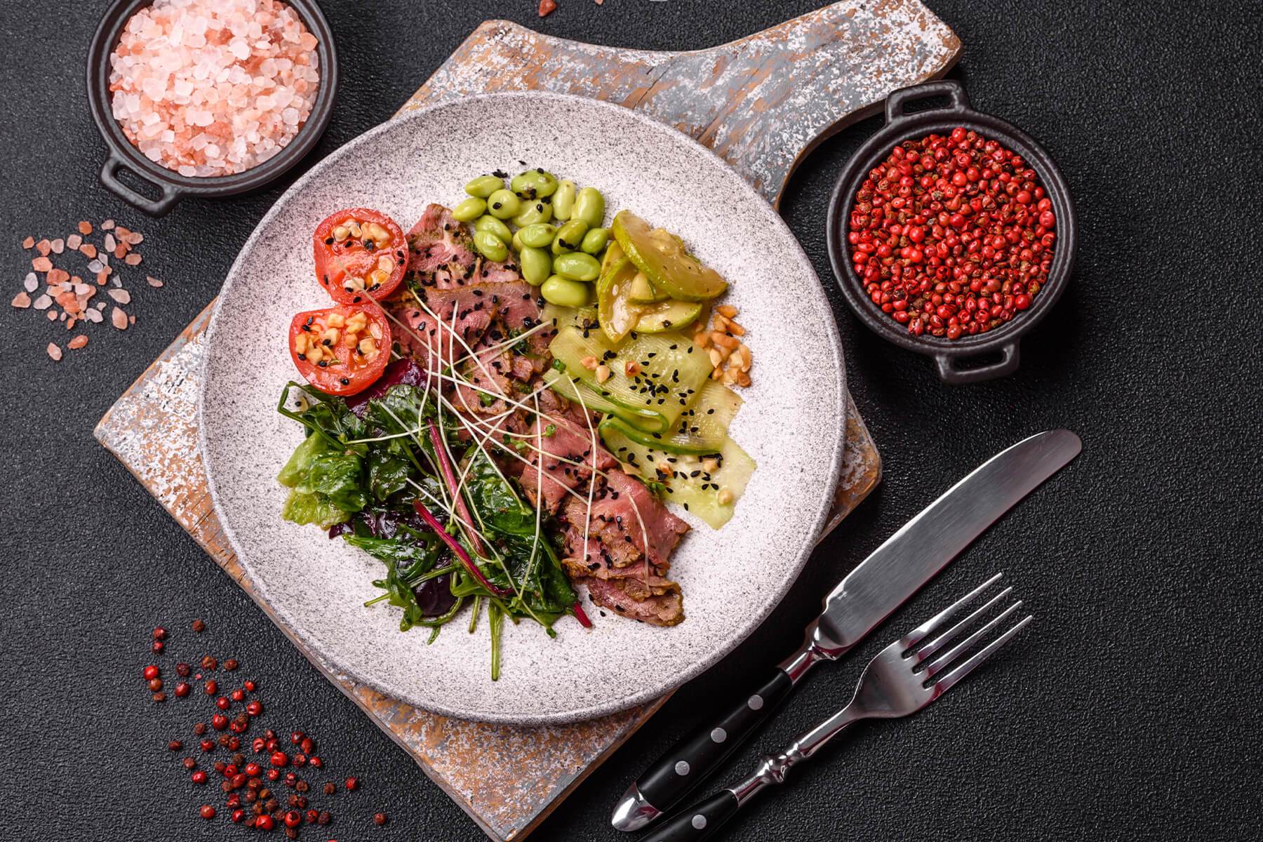 Warmer Roastbeef-Salat von oben auf einem Holzbrett, mit geschwenkten Blattsalaten, Edamame, warmen Gurkenbändern und Tomatenhälften, daneben Salz und rosa Pfeffer