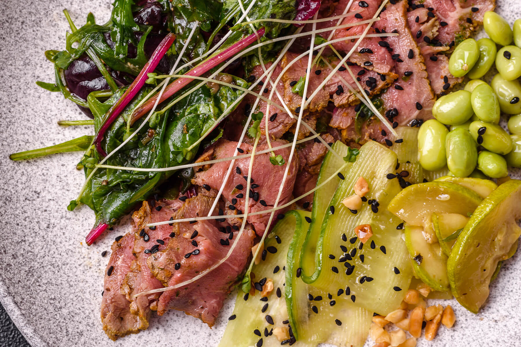Nahaufnahme von warmem Roastbeef-Salat mit rosa Rindfleischscheiben, Edamame, glasigen Gurkenbändern, schwarzem Sesam und feinen Sprossen