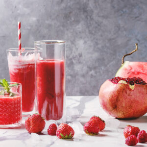 Drei Gläser roter Frucht-Smoothie mit Wassermelone und Beeren, daneben eine halbe Wassermelone und ein Granatapfel