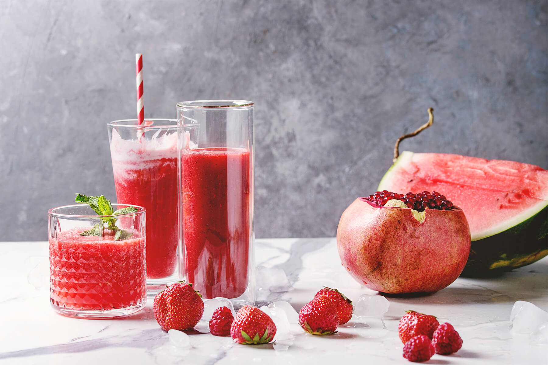 Drei Gläser roter Frucht-Smoothie mit Wassermelone und Beeren, daneben eine halbe Wassermelone und ein aufgebrochener Granatapfel