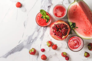 Roter Frucht-Smoothie in Gläsern von oben, garniert mit Granatapfelkernen und Minze, daneben Wassermelone und Erdbeeren