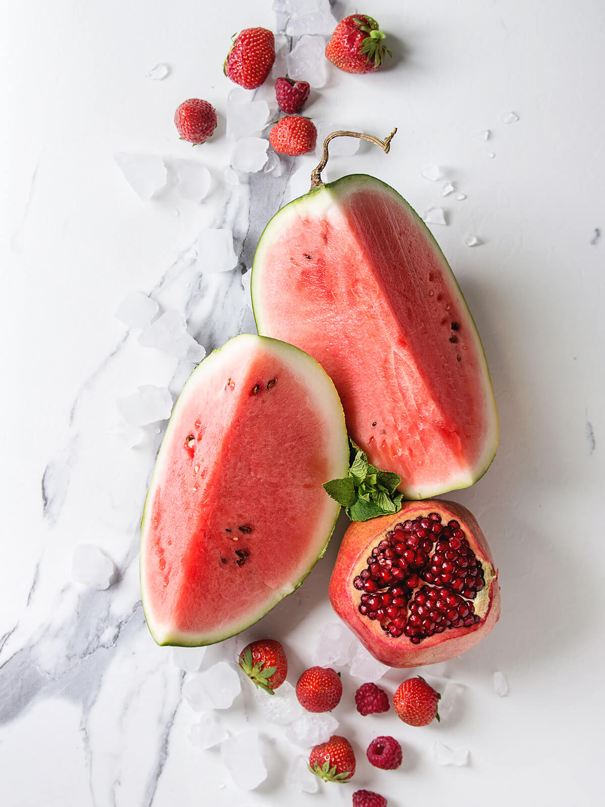 Zutaten für den roten Frucht-Smoothie – aufgeschnittene Wassermelone, Erdbeeren, Himbeeren, Granatapfel und Eis auf Marmor