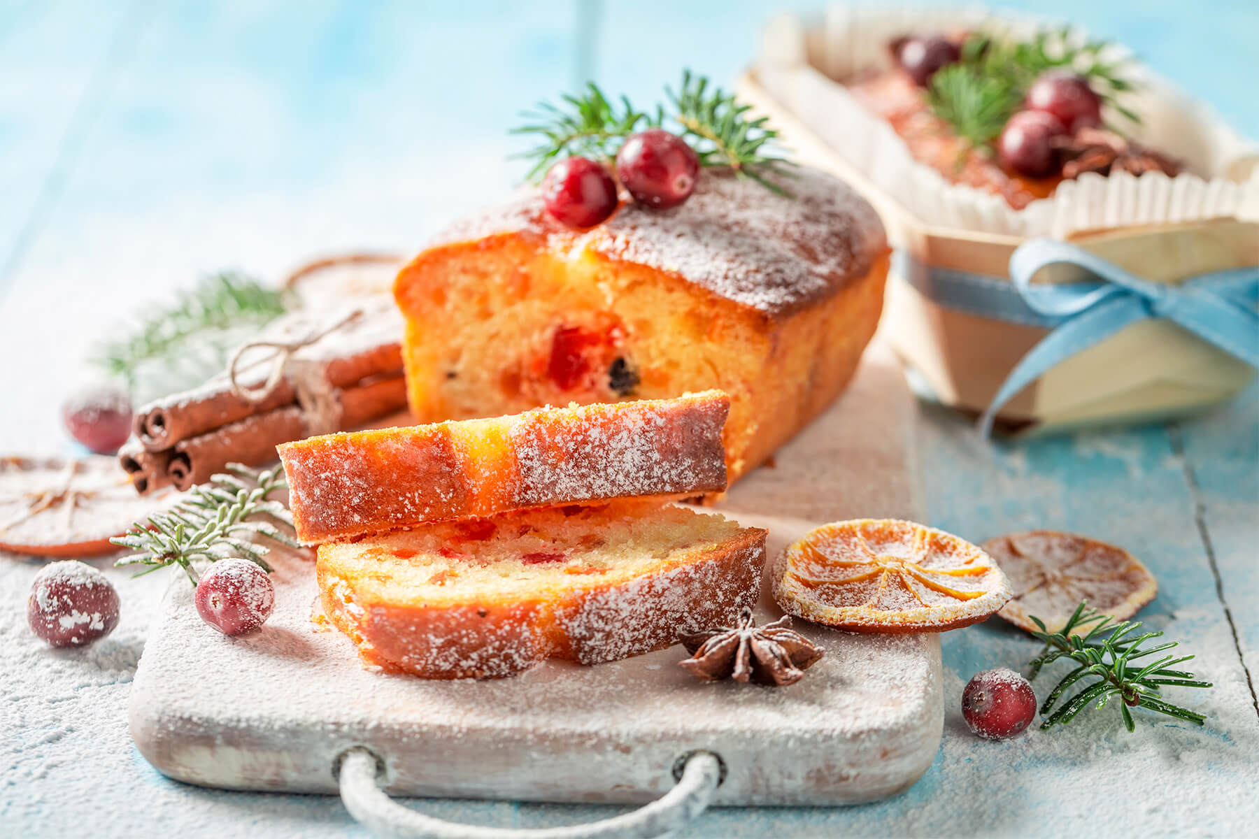 Angeschnittener weihnachtlicher Früchtekuchen mit roten kandierten Kirschen und Orangeat im goldgelben Teig, daneben Zimtstangen und getrocknete Orange