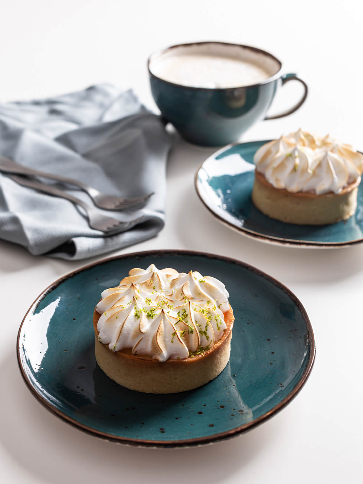 Zwei Mini-Zitronen-Baiser-Tartelettes mit knusprigem Mürbeteig, Zitronencreme und goldbraun abgeflämmter Baiserhaube auf petrolfarbenen Tellern, dahinter eine Tasse Milchkaffee