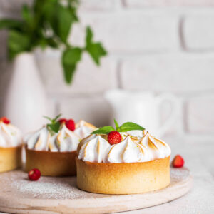Drei Mini-Zitronen-Baiser-Tartelettes mit knusprigem Mürbeteig, Zitronencreme und goldbraun abgeflämmter Baiserhaube auf einem hellen Holzbrett, garniert mit Walderdbeeren und Minze