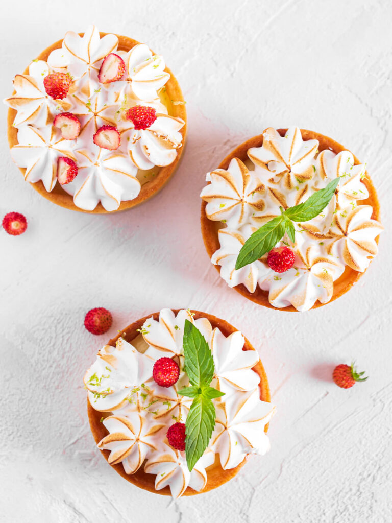 Drei fertige Mini-Zitronen-Baiser-Tartelettes von oben mit goldbraun abgeflämmter Baiserhaube, Limettenschale, Minze und Walderdbeeren