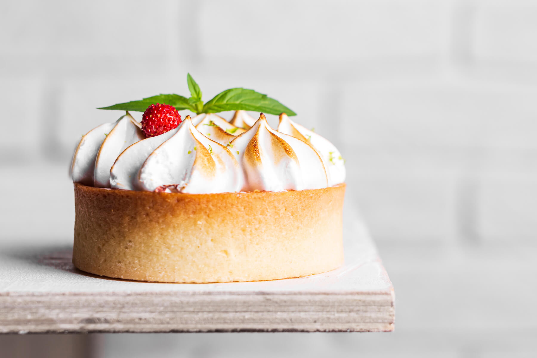 Seitenansicht eines Zitronen-Baiser-Tartelettes mit hohem Mürbeteigrand und aufgespritzten Baisertropfen, an Spitzen und Kanten goldbraun abgeflämmt, garniert mit Minze und einer Walderdbeere