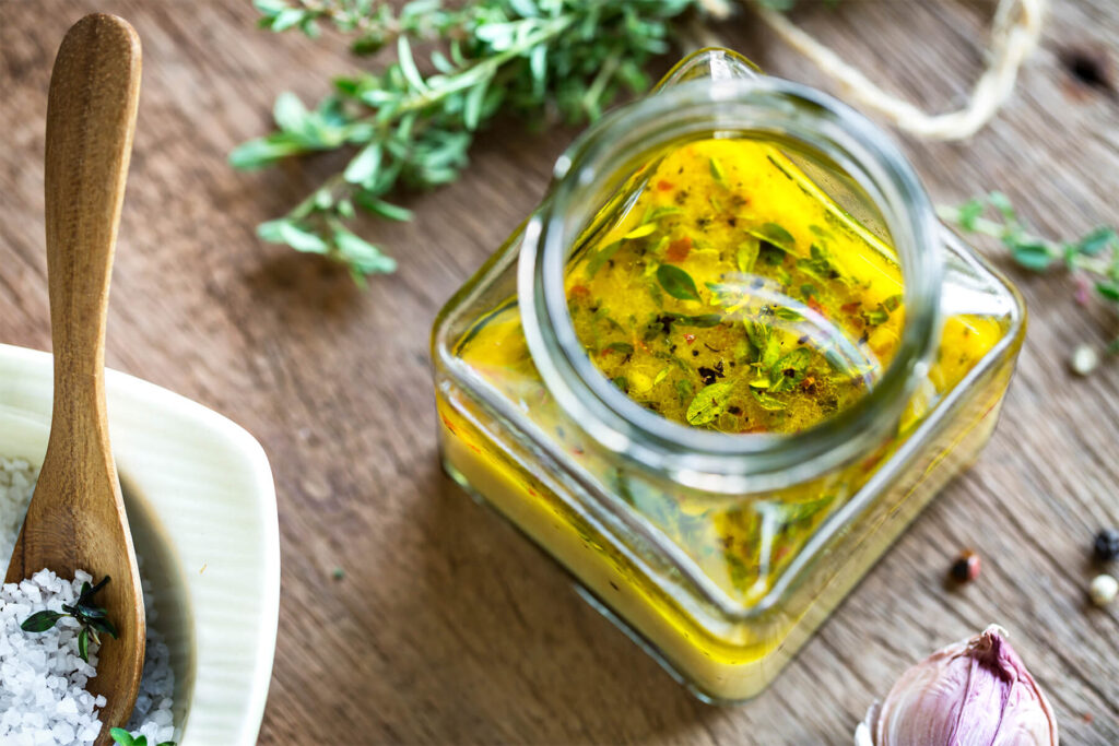 Zitronen-Thymian-Vinaigrette in einem offenen eckigen Glas mit schwimmenden Thymianblättchen und bunten Pfefferkörnern