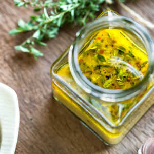 Zitronen-Thymian-Vinaigrette in einem offenen eckigen Glas mit schwimmenden Thymianblättchen und bunten Pfefferkörnern