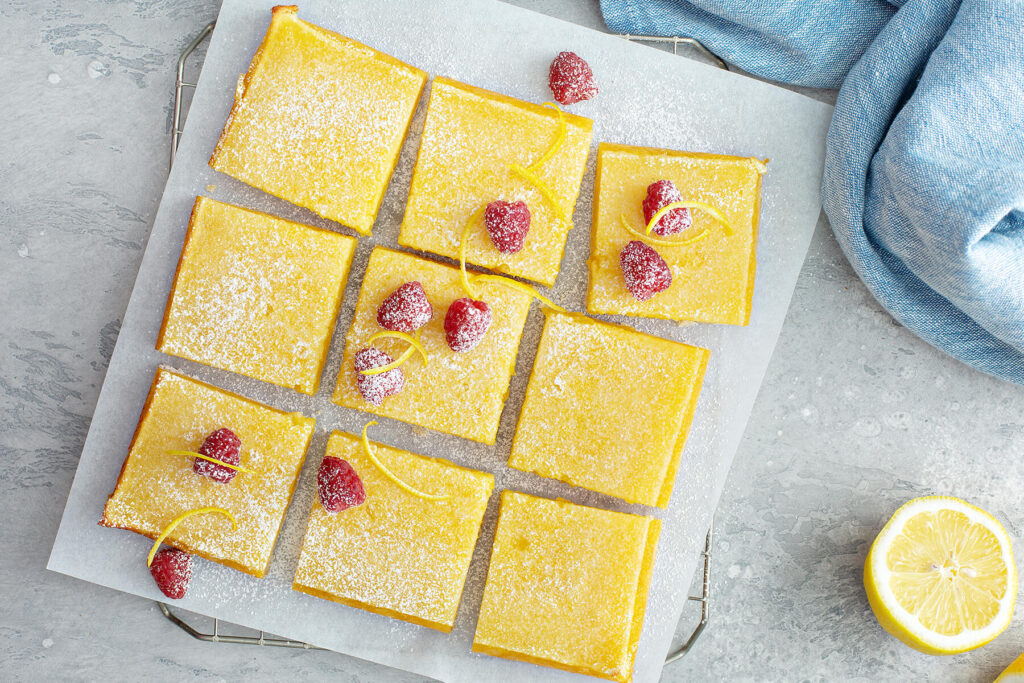 Aufsicht auf neun goldgelbe Zitronenschnitten auf Backpapier auf einem Kuchengitter, mit Puderzucker bestäubt und mit frischen Himbeeren und feinen Zitronenzesten-Spiralen garniert, daneben eine halbe Zitrone auf hellem Untergrund