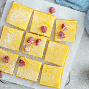 Aufsicht auf neun goldgelbe Zitronenschnitten auf Backpapier auf einem Kuchengitter, mit Puderzucker bestäubt und mit frischen Himbeeren und feinen Zitronenzesten-Spiralen garniert, daneben eine halbe Zitrone auf hellem Untergrund